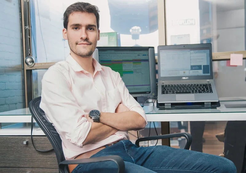 Junger Mann im modernen Büro sitzt entspannt vor Laptop und Monitor mit Business-Software.