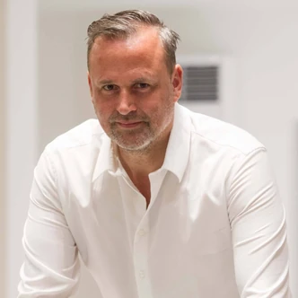 Mann mit weißem Hemd blickt freundlich in die Kamera vor neutralem Hintergrund.